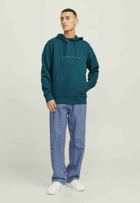 Sweat à capuche teal avec cordons et poche avant, featuring texte du logo blanc. Associé à un jean bleu clair et des baskets blanches. Design simple.