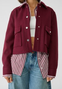 Burgunderrød uldblandingsjakke med trykknapper, forsynet med to forreste lommer og et cropped design, lagt over en hvid og rød stribet skjorte.