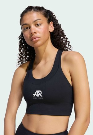 Donna con capelli ricci che indossa un reggiseno sportivo nero Adidas Runners, in piedi davanti a uno sfondo chiaro uniforme.