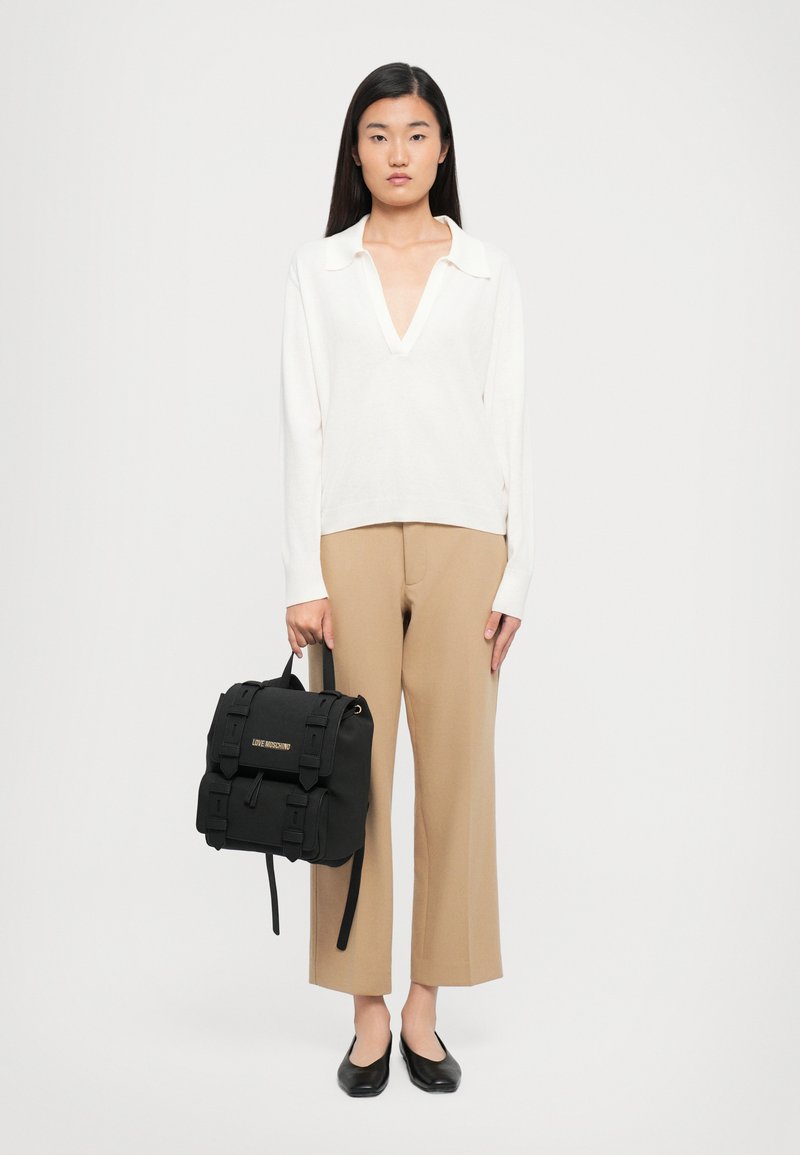 Γυναίκα με λευκή μπλούζα με μακρύ μανίκι και V-ντεκολτέ, και μπεζ κοντά παντελόνια, κρατώντας μια μαύρη τσάντα πλάτης Love Moschino, φορώντας μαύρες μπαλαρίνες σε λευκό φόντο.