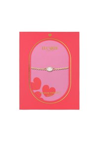 Pulsera dorada con un centro de piedra ovalada, presentada sobre un fondo rosa en un embalaje con patrón de corazones rojos. El texto dice "Acero Inoxidable Premium".