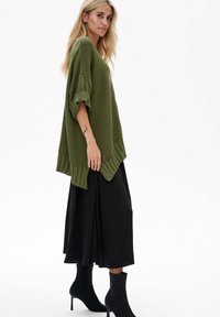 Przesadny zielony sweter z ribbowanym splotem i podwiniętymi rękawami, w połączeniu z lejącą czarną spódnicą maxi i czarnymi botkami za kostkę.