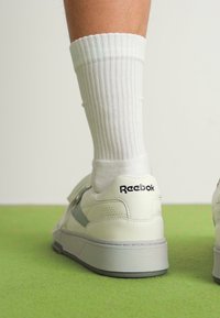 White leather sneakers with a grey accent, featuring a textured sole and Reebok branding on the heel, paired with ribbed white socks.