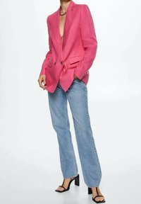 Femme portant un blazer rose vif, un jean droit bleu clair et des sandales à talons noirs, debout avec une main dans la poche.