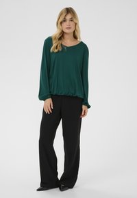 Zelena bluza dugih rukava s zaobljenim izrezom i nabranim rubom, uparen s crnim hlačama širokih nogavica. Tkanina izgleda mekano i lepršavo.