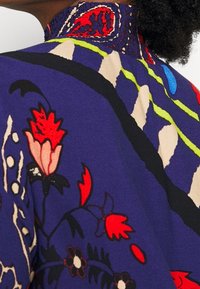 Blauwe stof met bloemendessin en details in rood, groen en crème; heeft een gesmockte kraag. Het ontwerp bevat bloemen en abstracte vormen.