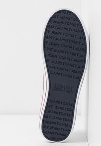 Czarna gumowa podeszwa sportowa z fakturowanym logo, z napisem "TOMMY JEANS". Podkreślona białą obwódką i cienką czerwoną linią wzdłuż krawędzi.