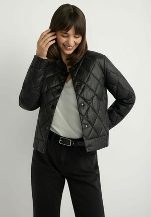 Czarna pikowana kurtka z wysokim kołnierzem, zatrzaskami i teksturowanymi detalami wzdłuż dołu, noszona na jasnej koszuli i czarnych spodniach.