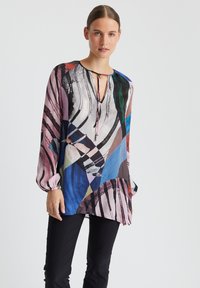 Blouse à manches longues avec un motif abstrait multicolore, en tissu léger, décolleté en V avec un lien, et manches bouffantes. Pantalons noirs en dessous.