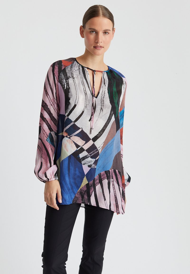 Blouse à manches longues avec un motif abstrait multicolore, en tissu léger, décolleté en V avec un lien, et manches bouffantes. Pantalons noirs en dessous.