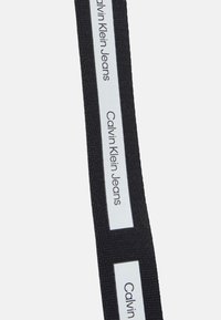 Černý textilní popruh se dvěma bílými obdélníkovými štítky, na kterých je černým písmem nápis "Calvin Klein Jeans". Odolná textura a čistý design.