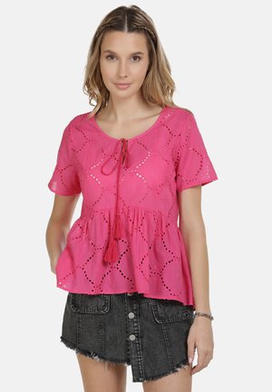 Donna che indossa una camicetta rosa brillante a maniche corte con motivi traforati e lacci con nappine, abbinata a una gonna di jeans nera con bottoni.