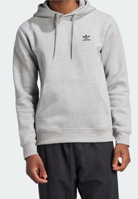 Grå huva sweatshirt tillverkad av mjukt tyg, med en framficka och en svart Adidas-logga på vänster bröst. Förstärkta manschetter och nederkant.
