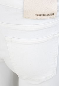 White denim jeans featuring a back pocket with subtle stitching details and a textured beige leather label with "True Religion" embossed.