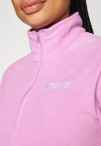 Lys rosa fleecejakke med høy krage, halvlukking med glidelås, og sølvfarget "Terrex"-logo på brystet. Myk tekstur og jevn overflate.