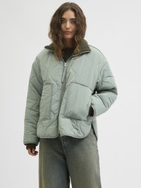 Ženska s kodrastimi lasmi nosi svetlozeleno puhasto jakno z velikimi sprednjimi žepi preko temnega puloverja in ohlapnih jeans hlač.