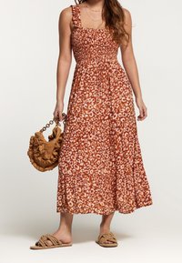 Robe léopard à imprimé rouge et sable avec corsage smocké, bretelles froncées et jupe à volants, associée à un sac à main texturé marron et des sandales en corde.