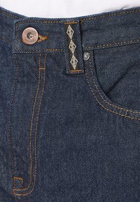 Jean en denim de couleur bleu foncé, avec des coutures orange contrastantes, un système de fermeture à bouton en métal et une étiquette en tissu noir brodée avec un logo.