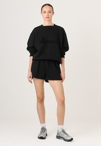 Čierna mikina s výšivkou "La marie", vo voľnom strihu, kombinovaná s čiernymi šortkami. Model má na sebe strieborné športové topánky a biele ponožky.