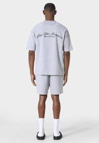 Man staat met zijn rug naar de camera, draagt een lichtgrijs oversized T-shirt en bijpassende shorts, witte sportsokken en zwarte slippers.