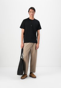 Tânăr stând în fața unui zid alb, îmbrăcat cu un tricou negru, pantaloni bej de tip wide-leg, pantofi maro, ținând o geantă mare de tip tote neagră în mâna stângă.