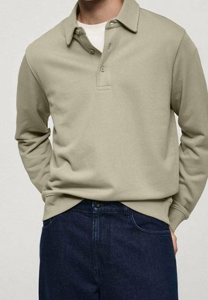 Hombre lleva una camisa tipo polo de manga larga beige sobre una camiseta interior blanca, combinada con jeans de mezclilla azul oscuro, de pie con las manos en los bolsillos.