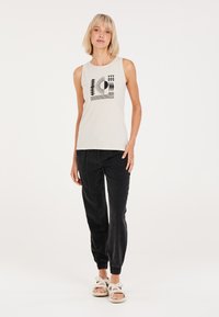 Top senza maniche crema con una grafica geometrica nera; abbinato a pantaloni jogger in corduroy neri e sandali bianchi con doppie cinghie.
