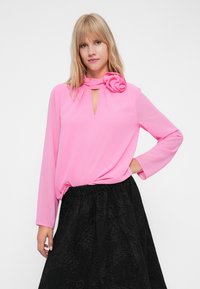 Rosa långärmad Blus med en knuten halsdetalj och blommig accent, tillsammans med en texturerad svart kjol med ett mönster.