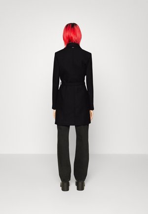 Personne aux cheveux rouge vif portant un manteau noir ceinturé, un pantalon noir et des chaussures plateau noires, debout face à un mur blanc.