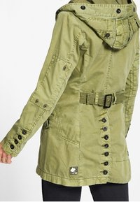 Olivgrön jacka i slitstarkt tyg; har en dragsko i huvan, bältad midja, stora knappar och flera fickor på ärmar och sidor.