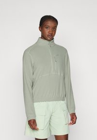 Light green half-zip fleece pullover with a high collar, featuring stitched detail and a small zippered chest pocket. Paired with light green shorts.
