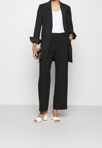 Blazer negro ajustado sobre una blusa blanca, combinado con pantalones de pierna ancha negros y sandalias de tacón bloque abiertas en la parte delantera blancas. Sostiene un bolso de mano negro.