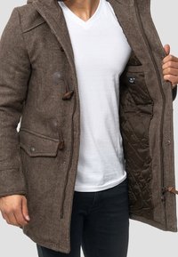 Mand iført en brun struktureret togglejakke, der viser sin quiltede indvendige foret, parret med en hvid V-hals skjorte og mørke bukser.