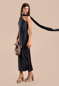 Schwarzes Satin-Halterkleid mit einem Schlitz an der Seite, kombiniert mit goldenen Plateausandalen und einer metallischen Clutch mit Schleifenakzent, die einen fließenden Rippenschmuck hat.