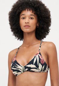 Blomster-bikini-topp med trekantet form, tynne justerbare stropper, og en mørk bakgrunn med grønne og rosa botaniske mønstre.