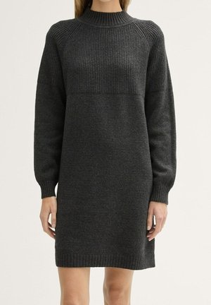Vrouw die een donkergrijze, langarmige gebreide sweaterjurk draagt met geribbelde boorden, zoom en hoge kraag, staand tegen een effen achtergrond.