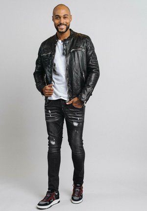 Veste en cuir noir avec des zips, t-shirt blanc, jeans noirs déchirés et baskets noires/rouges. Pose debout avec un arrière-plan neutre.