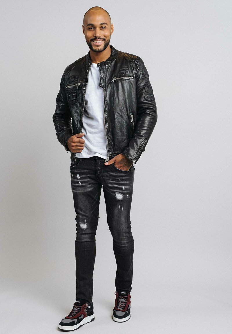 Veste en cuir noir avec des zips, t-shirt blanc, jeans noirs déchirés et baskets noires/rouges. Pose debout avec un arrière-plan neutre.