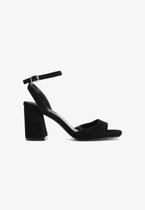 Mustat mokkanahkaiset korkokengät, joissa on paksu korko, avoin varvas ja säädettävä nilkkahihna. Suunnittelu on sileä ja minimalistinen.