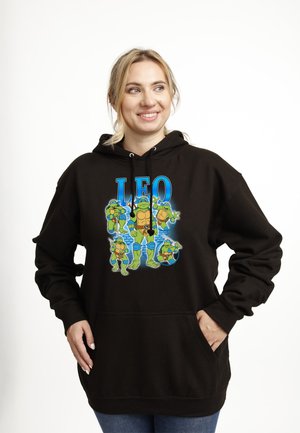 Smilende kvinde iført sort hættetrøje med fem Teenage Mutant Ninja Turtles-figurer og teksten "LEO" ovenover på hvid baggrund.