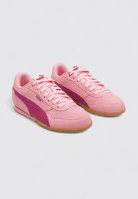 Zapatillas Puma en ante rosa con acentos en un tono de rosa más oscuro. Cuentan con suela de goma, puntera redonda y diseño con cordones. Logo dorado en la lengüeta.
