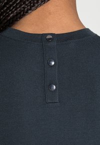 Dark teal knit top featuring a rounded neckline, visible ribbing, and three metallic snap buttons at the back for closure.