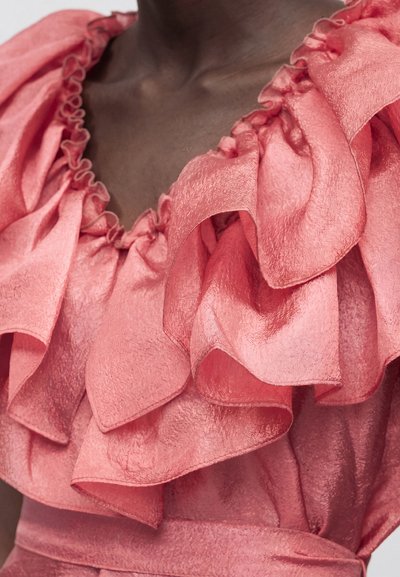 Κοντινό πλάνο ενός σκοτεινής επιδερμίδας ατόμου που φοράει ένα κοραλλί ροζ υφασμάτινο φόρεμα με μεγάλα πολυεπίπεδα βολάν γύρω από το λαιμό.