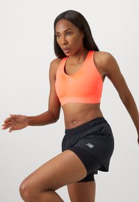 Mujer atlética con sujetador deportivo color coral y pantalones cortos de correr negros en medio de un movimiento, demostrando un entrenamiento dinámico sobre un fondo liso.