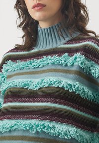 Monivärinen neulepaita, jossa on korkea resorikaulus. Päällä vaakasuoria raitoja sävyissä tehostevihreä, viininpunainen ja oliivinvihreä, teksturoiduilla hapsuilla koristeltuna.