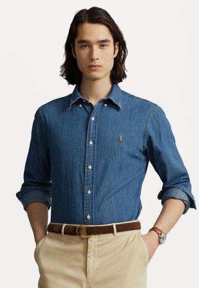 Denim button-up shirt in medium blue with long sleeves, rolled at the cuffs, features a small logo and white buttons, paired with beige trousers.