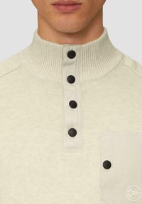 Lichtbeige pullover met een geribde hoge kraag, voorzien van zwarte knoopaccenten en een textuur gebreid patroon, inclusief een borstzak.