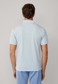 Man med kort brunt hår iklädd en ljusblå kortärmad polo och blå byxor står vänd bort från kameran mot en neutral bakgrund.