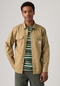 Tan button-up shirt met twee voorzakken, gedragen over een groen-wit gestreept t-shirt. Model heeft kort haar en draagt groene broek.