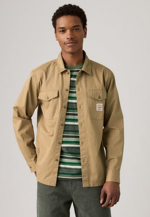 Tan button-up shirt with two front pockets, worn over a green and white striped t-shirt. Model has short hair and is wearing green pants.
